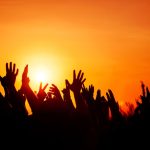 An image of silhouetted hands raised up to the sky and setting sun.