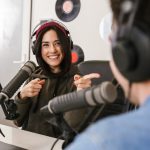 A smiling podcaster points to her guest with his back to the camera
