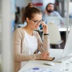 A reporter works at her desk, taking notes for a story while talking with a source on the phone.