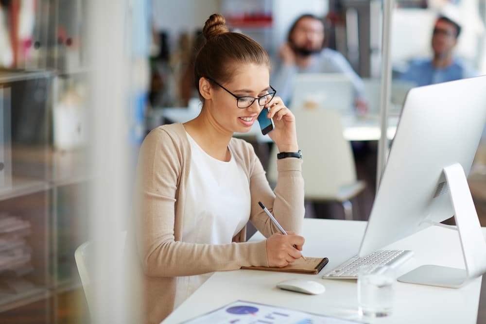 A reporter works at her desk, taking notes for a story while talking with a source on the phone.
