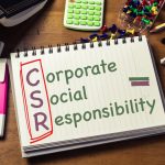 A close-up image of a desk with various work items and a notebook with the words Corporate Social Responsibility in the center.