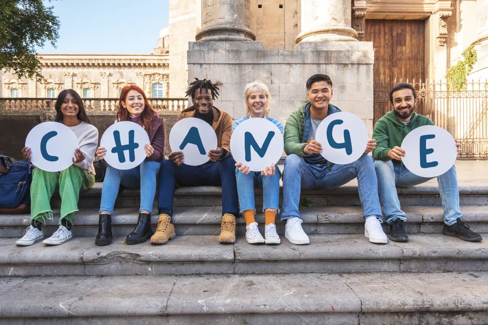 A diverse group of college students sitting outside, each holding a single letter sign that together spells the word “change.”